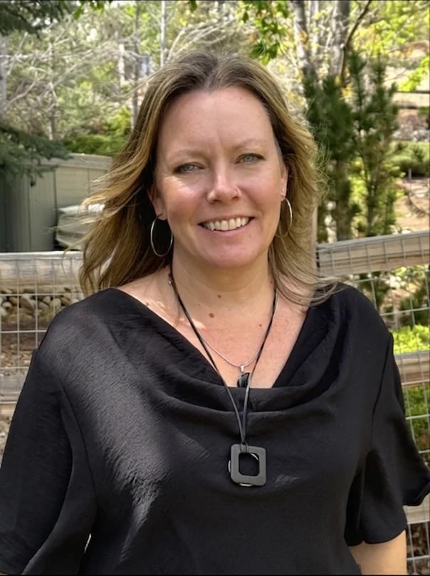 Woman with brown wavy hair smiling at camera, wearing black short-sleeved top with pendant necklace, garden setting background