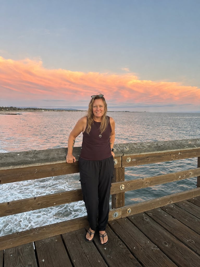 Woman standing on wooden pier at sunset with orange-tinted clouds and ocean waves in background